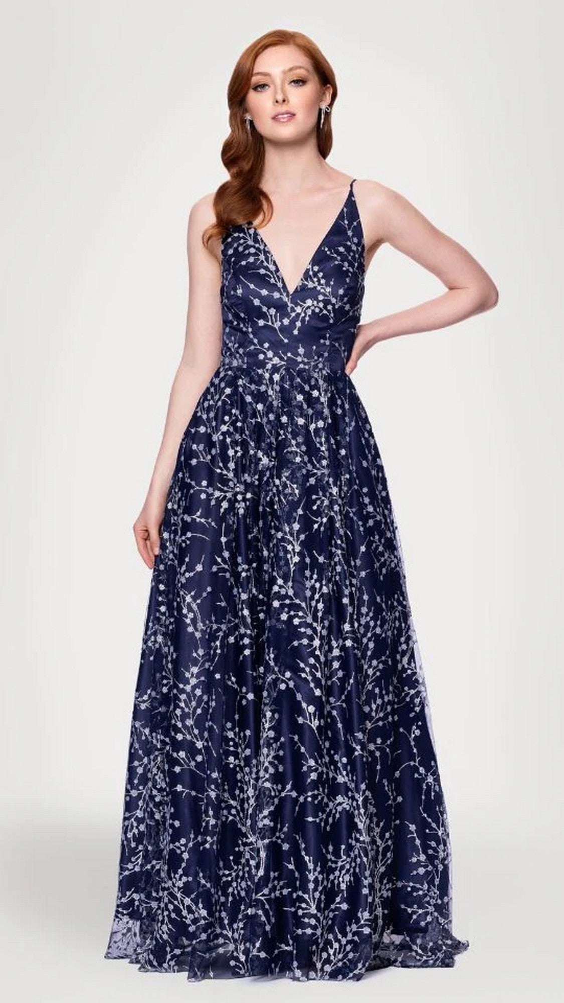A woman stands with hand on hip in the Blondie Nites 5386BN long prom dress, featuring a sleeveless, deep V-neck navy/peri design with a white floral pattern and an elegant A-line skirt, against a plain light gray background.